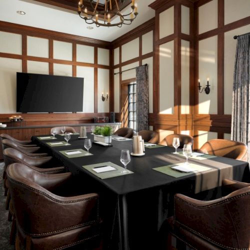 A formal, wood-paneled conference room with a long black table, brown leather chairs, a wall-mounted TV, and large windows with curtains.