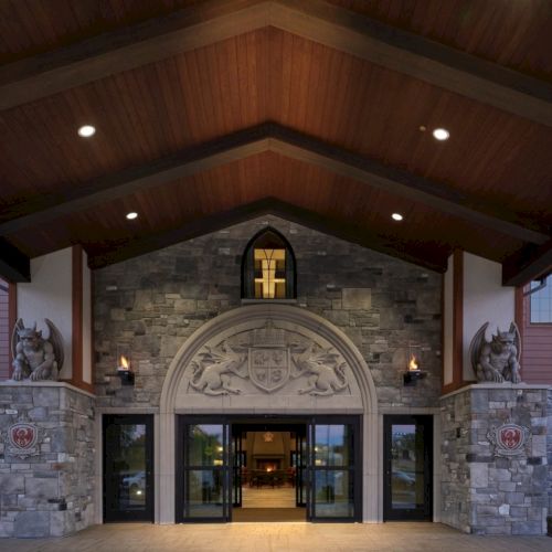 An entrance to a stone building with a wooden peaked roof, grand double doors, stone lions, and a cross emblem above, warmly lit.