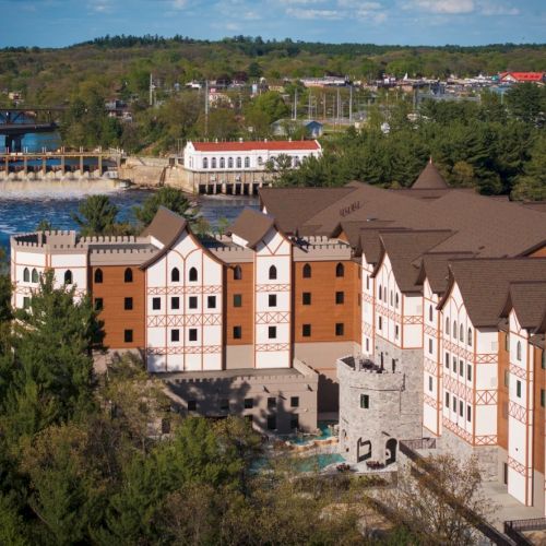 A scenic resort with castle-like buildings along a forested shoreline, overlooking a lake and distant hills, evoking a European-inspired riverside village.