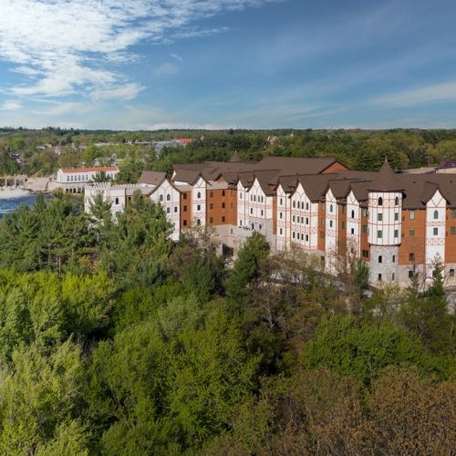 A row of multi-story beige and orange apartment buildings sits along a lush green hillside under a blue sky, with a lake to the left.