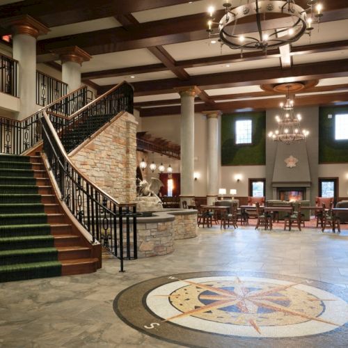 A grand hotel lobby with a curved staircase, marble floors, chandeliers, round compass emblem on the floor, and seating lounge area.