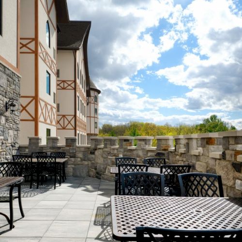 A stone-and-wood terrace with metal lattice tables and chairs, a stone railing, and a building with half-timbered walls; sunny, partly cloudy skies.