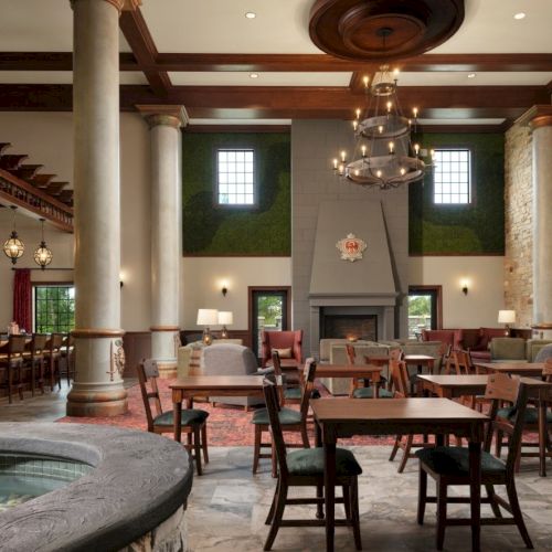 A stylish, spacious dining hall with high ceilings, stone walls, and a central fireplace; multiple wooden tables and chairs create a warm, inviting dining area.