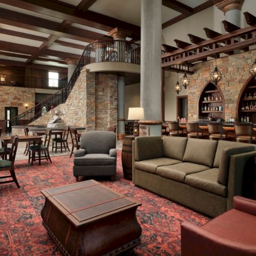 A cozy, upscale hotel lobby with a stone bar, arched shelves, warm lighting, a red patterned rug, and comfortable seating around wooden tables.