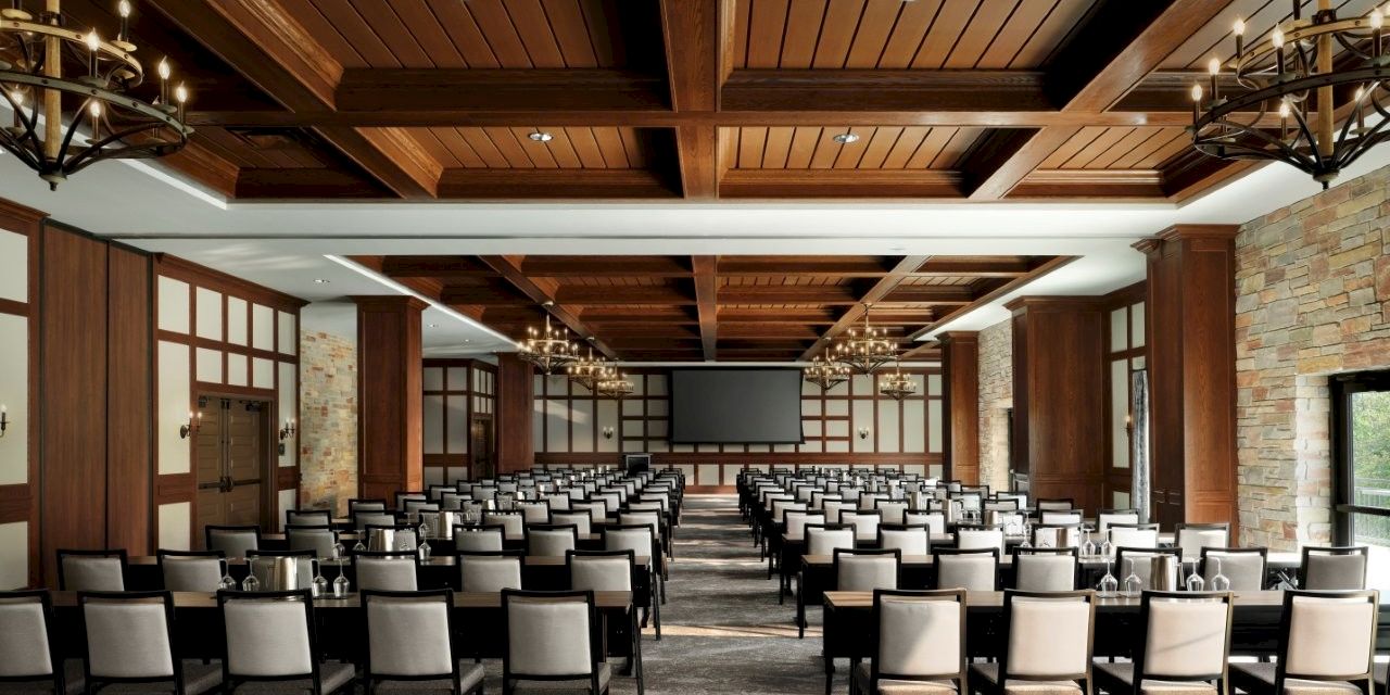A large conference room set up for a presentation, with rows of chairs facing a screen at the front, chandeliers, and wood paneling, all in one spacious hall.