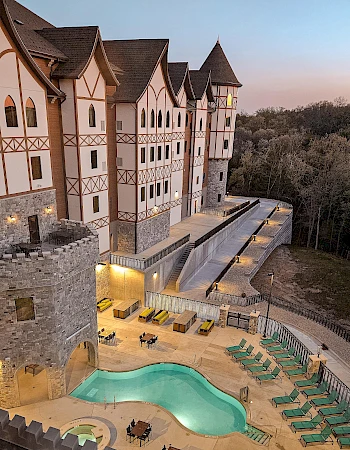 A medieval-style hotel with stone towers overlooks a curvy pool and loungers, set amid wooded hills at dusk.