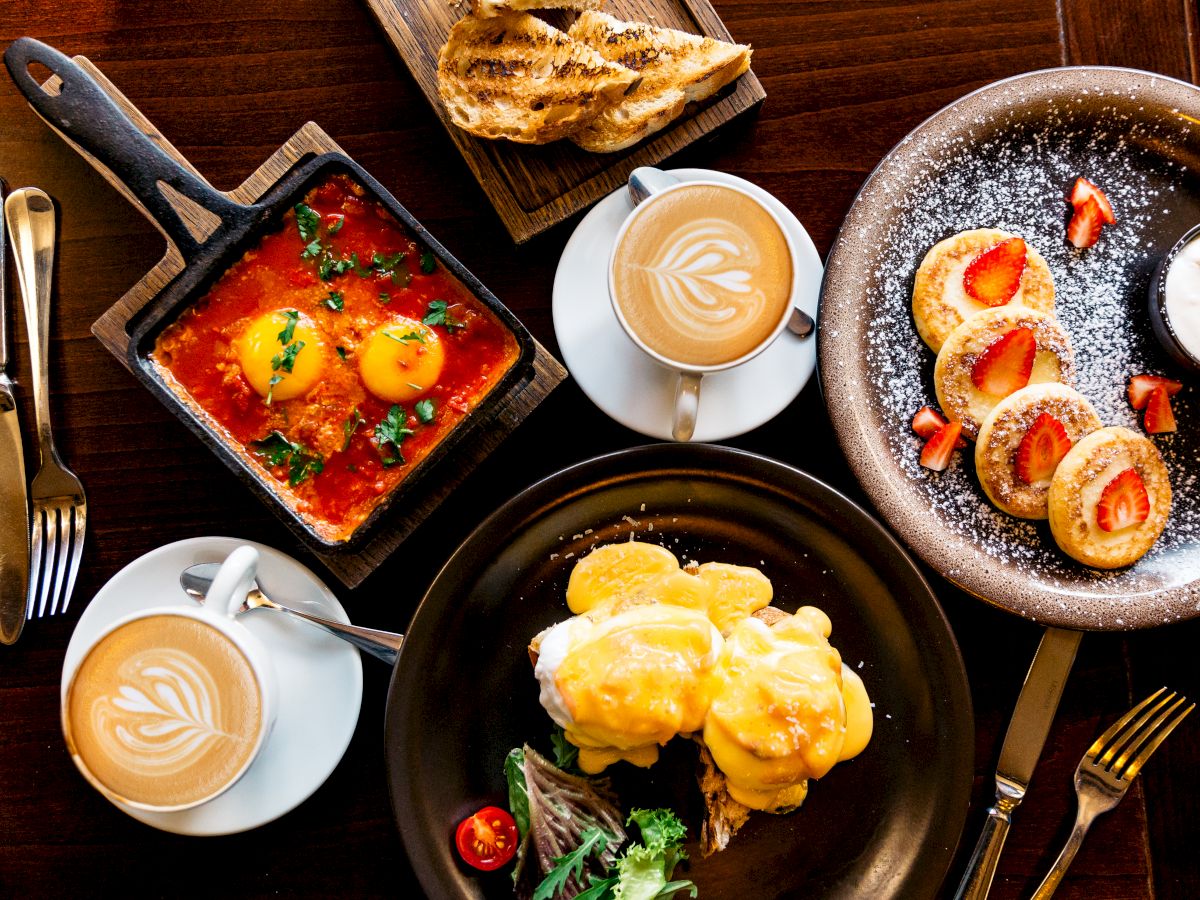 A top-down breakfast spread with shakshuka, croissants, rainbow waffles, and a cappuccino, plus a plate of eggs Benedict and herbs.