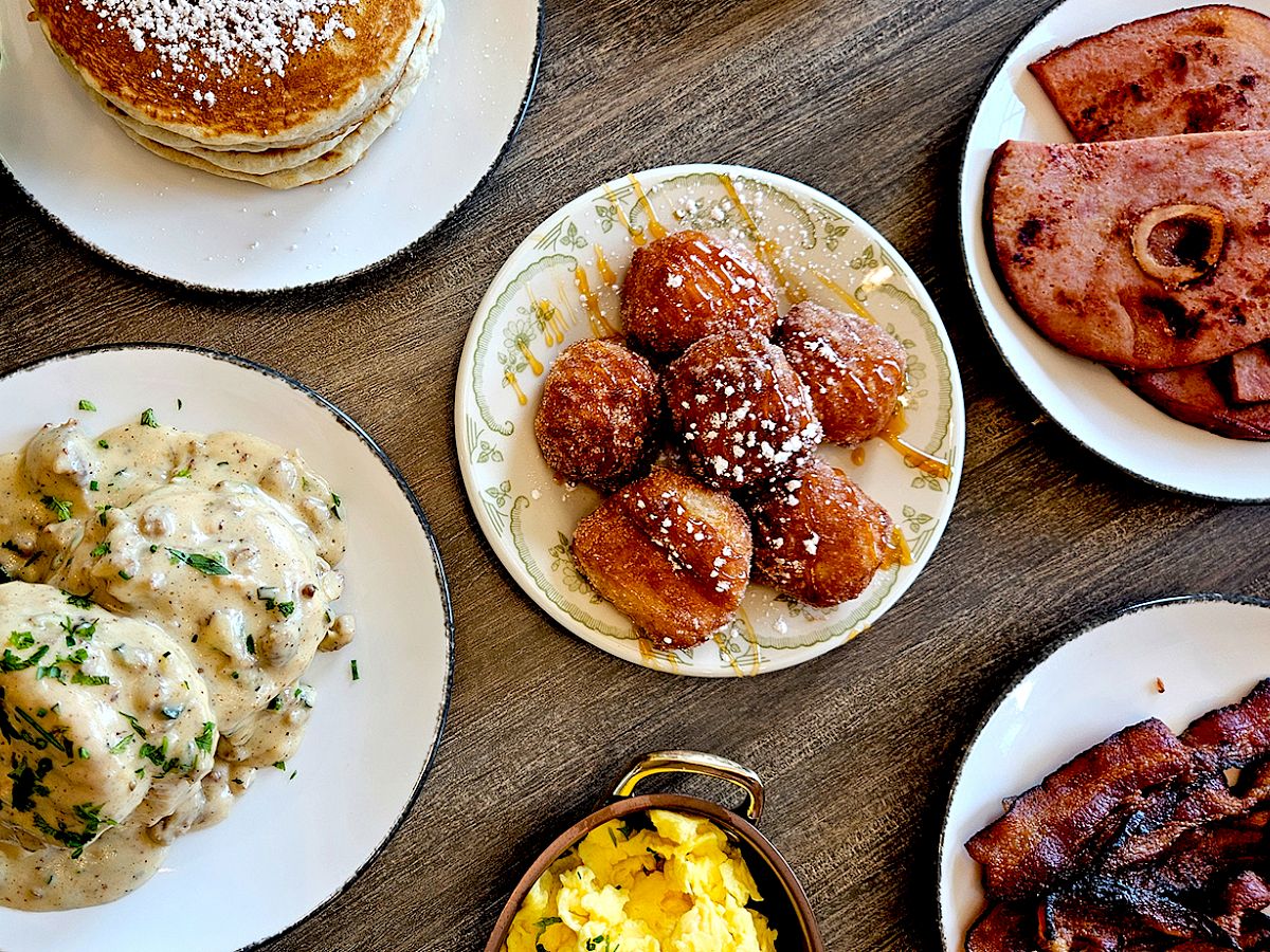 Plates of comfort food&mdash;creamy chicken, glazed fried dough balls, and cheesy pizza slices&mdash;set on a wooden table.