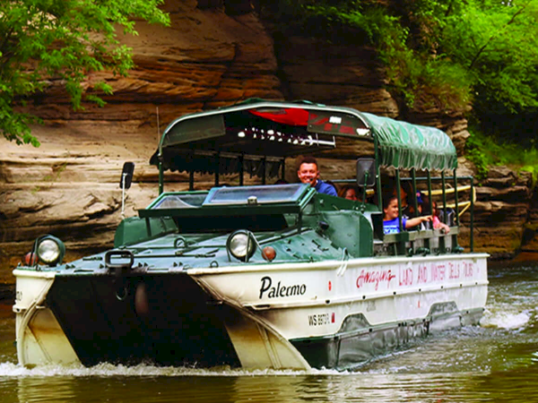 A pontoon tour boat with a green canopy sails on a river, passing a sandstone cliff; passengers enjoy the ride along lush greenery.