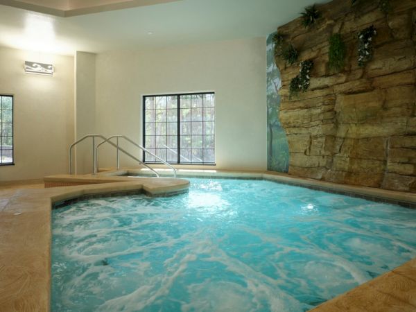 A cozy indoor pool area with a stone wall, bubbling turquoise water, and metal handrails by the steps, under warm lighting.