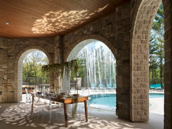 A stone patio with arches overlooks a pool; sunlight creates dappled patterns on the floor near a wooden table and chairs.