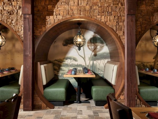 A cozy, booth-style restaurant nook with curved wooden partitions, warm lighting, green and beige seating, and a mural of trees in the background.