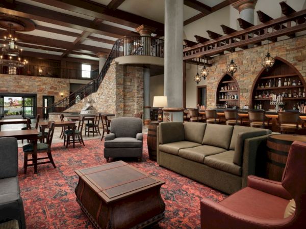 A cozy, upscale lounge with plush seating, a rich red carpet, stone walls, a wooden bar, and a mezzanine balcony overlook, inviting relaxation.