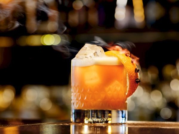 A rocks glass with an amber cocktail, large ice cube, an orange slice garnish, set against a blurred bar backdrop.