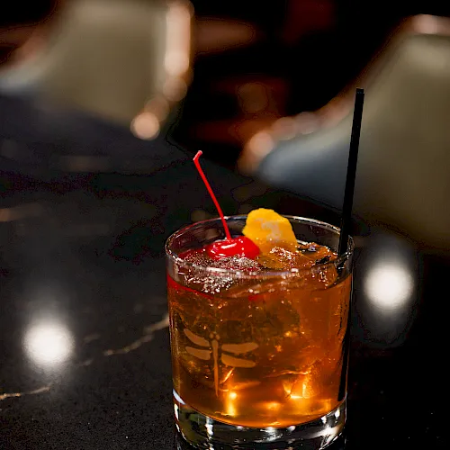 A whiskey cocktail on a dark bar, with ice, a red straw, a black straw, and a lemon twist garnish in a short glass. End.