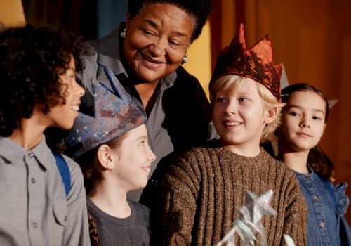 A group of kids wearing paper crowns smile together at a small party, guided by an adult, as they celebrate with a wand and festive vibes.