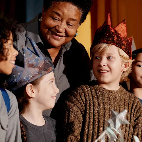 A group of kids wearing paper crowns smile together at a small party, guided by an adult, as they celebrate with a wand and festive vibes.