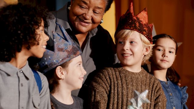 A group of kids wearing paper crowns smile together at a small party, guided by an adult, as they celebrate with a wand and festive vibes.