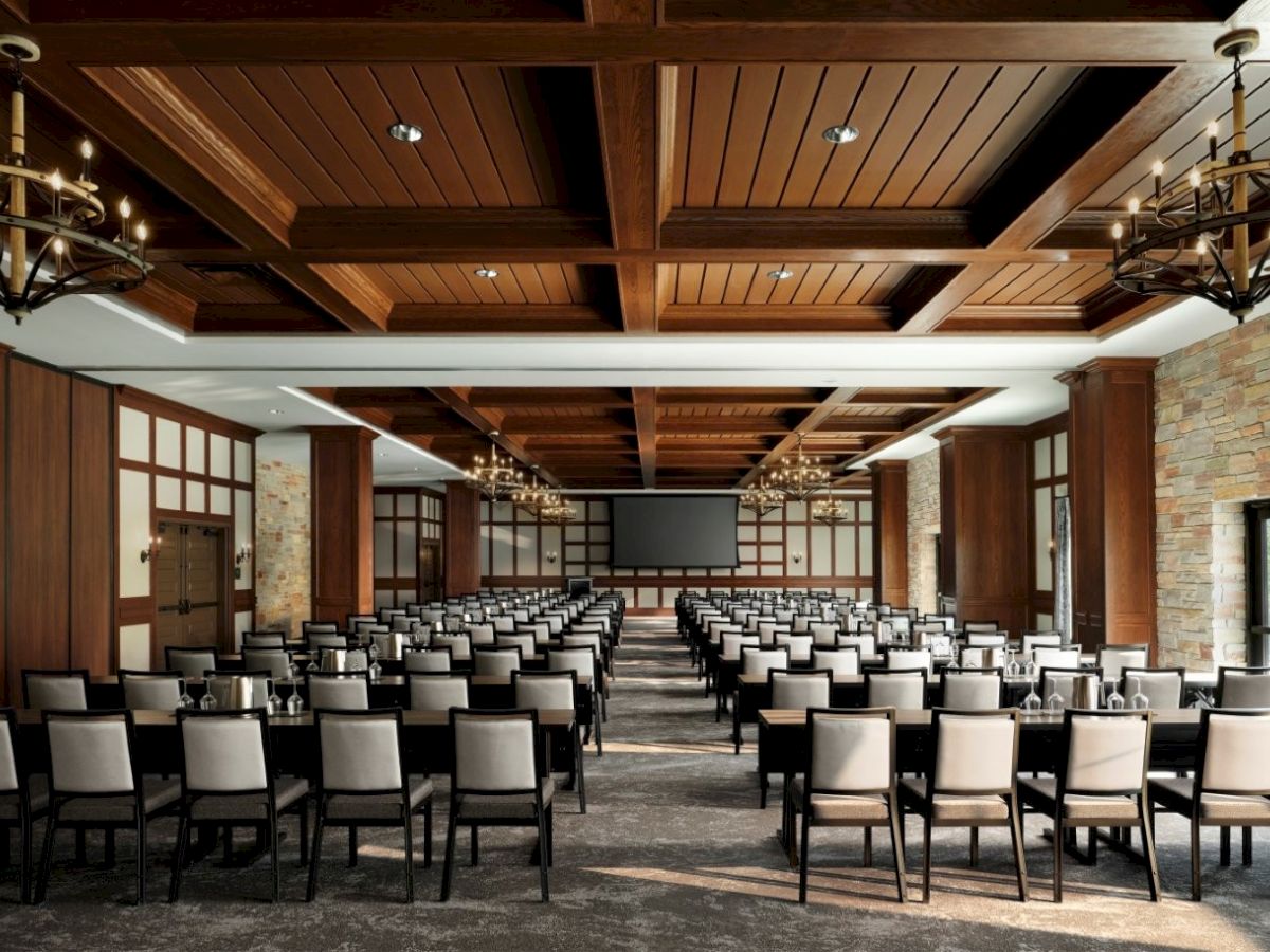 A spacious conference room with rows of chairs facing a large screen, chandeliers, wooden ceiling panels, and stone accents, set for a lecture.