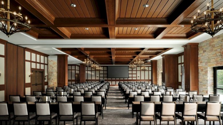 A spacious conference room with rows of chairs facing a large screen, chandeliers, wooden ceiling panels, and stone accents, set for a lecture.