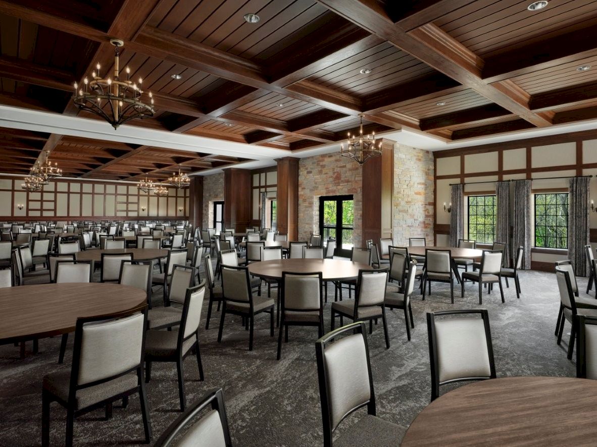 A spacious banquet hall with round and rectangular tables, upholstered chairs, chandeliers, wood coffered ceiling, and large windows, ready for dining.