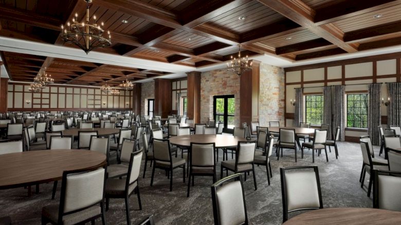 A spacious banquet hall with round and rectangular tables, upholstered chairs, chandeliers, wood coffered ceiling, and large windows, ready for dining.