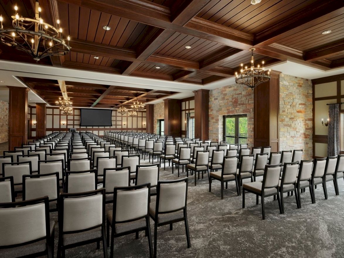 A spacious event hall with rows of black-and-white chairs, chandeliers, stone walls, and a projector screen.