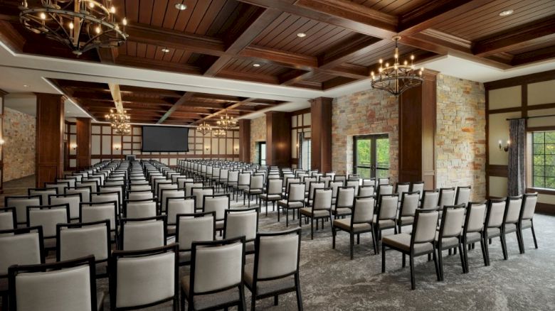 A spacious event hall with rows of black-and-white chairs, chandeliers, stone walls, and a projector screen.