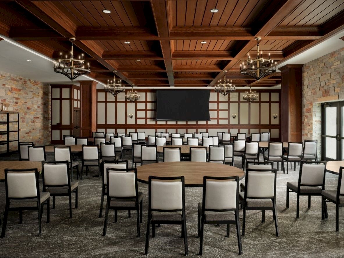A conference-style room with brick walls, wooden beam ceiling, multiple chandeliers, and rows of gray-upholstered chairs facing a large central screen.