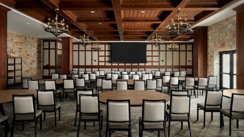 A conference-style room with brick walls, wooden beam ceiling, multiple chandeliers, and rows of gray-upholstered chairs facing a large central screen.