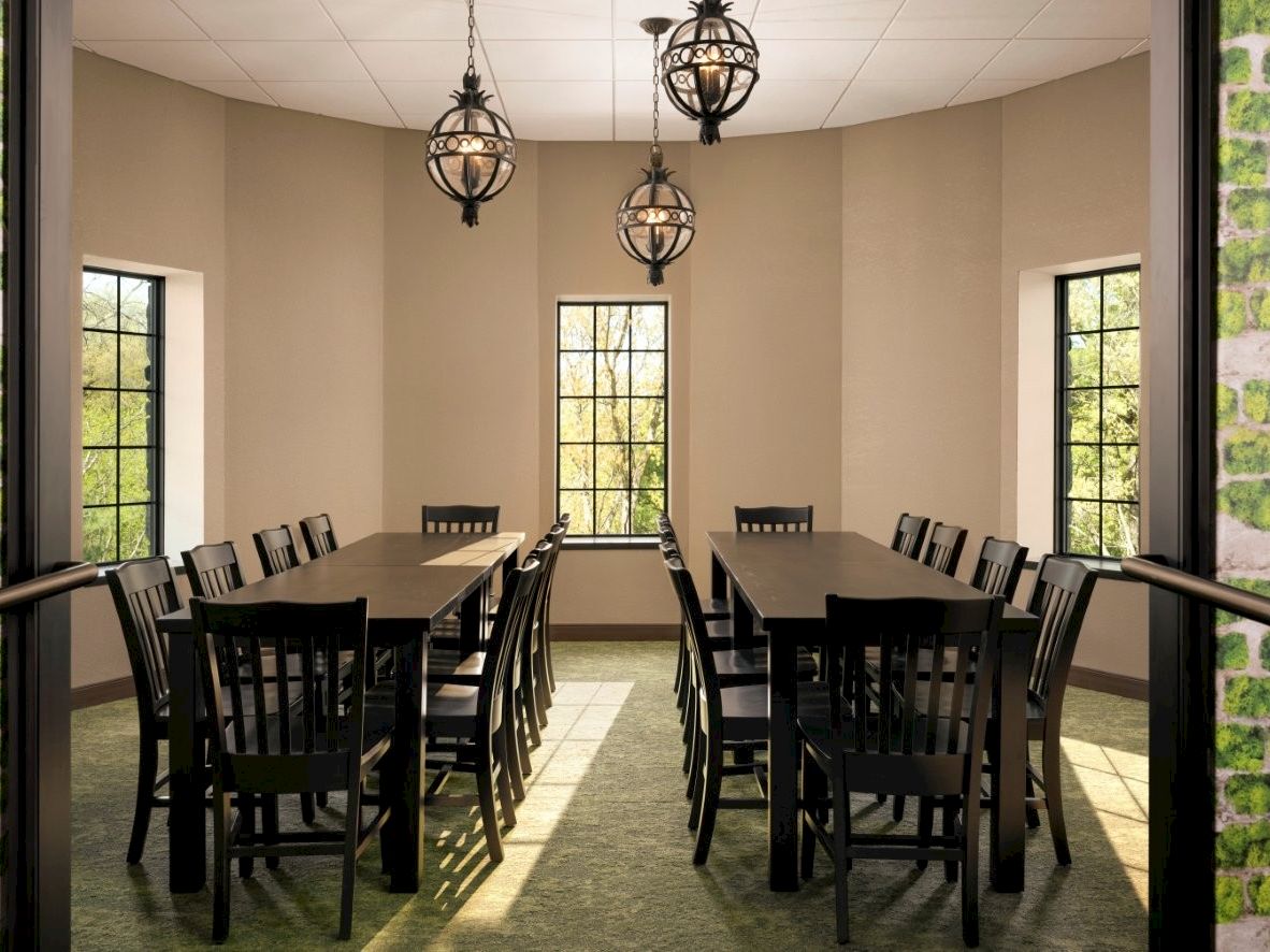 A bright dining room with two long dark tables, multiple black chairs, and three modern pendant lights hanging from a beige ceiling.