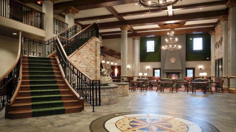 An elegant indoor hall with grand green-carpeted stairs, stone columns, chandeliers, a fireplace, chairs, and a star compass mosaic floor.