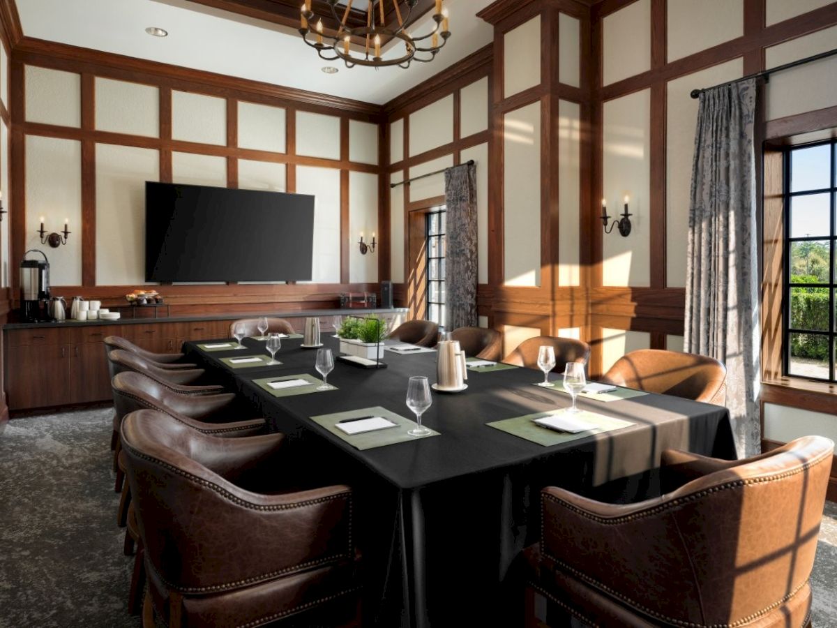 A formal meeting room with a long black-cloth table, brown leather chairs, wood-paneled walls, a large TV, and windows letting in natural light.
