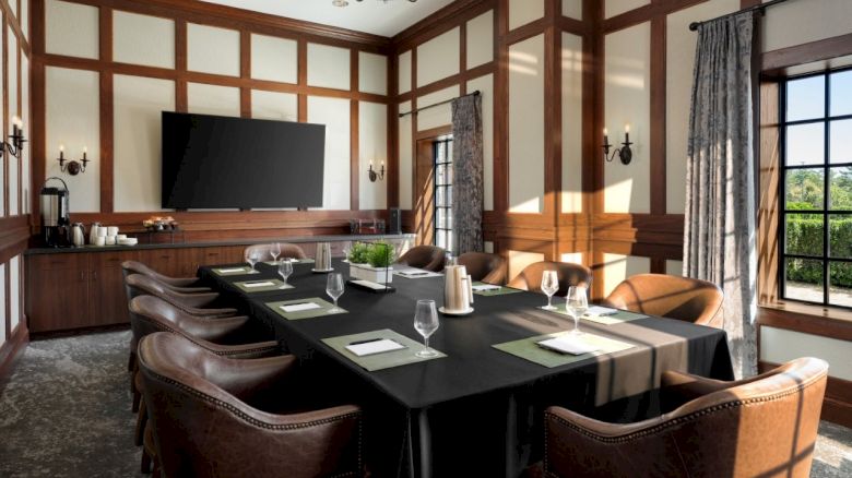 A formal meeting room with a long black-cloth table, brown leather chairs, wood-paneled walls, a large TV, and windows letting in natural light.