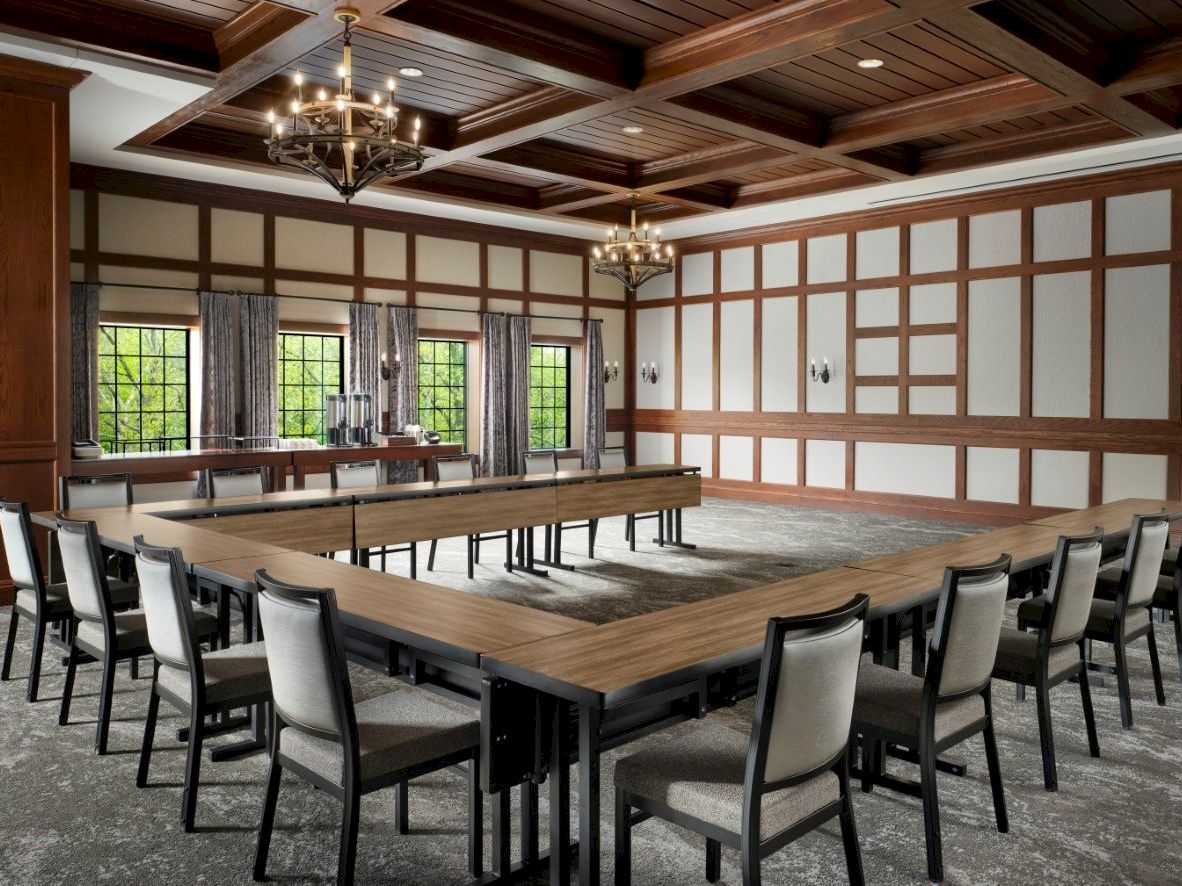 A large banquet hall with a long wooden table, matching chairs, chandeliers, and tall windows in a timber-paneled room.