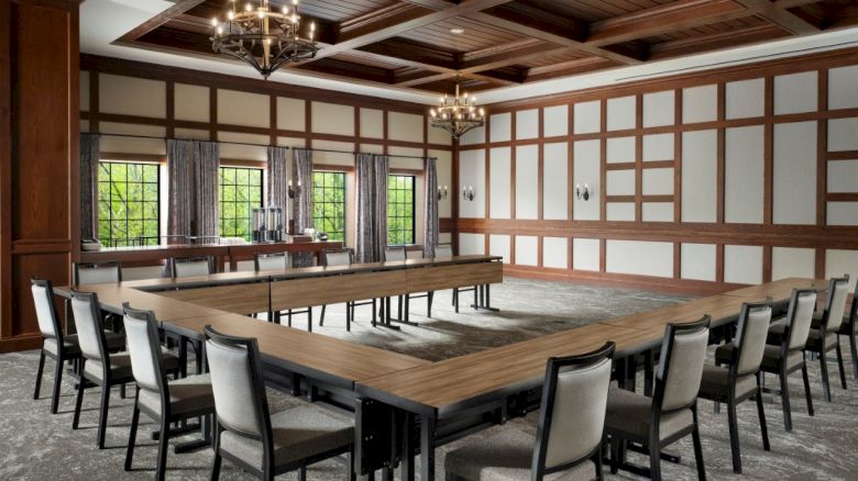 A large banquet hall with a long wooden table, matching chairs, chandeliers, and tall windows in a timber-paneled room.