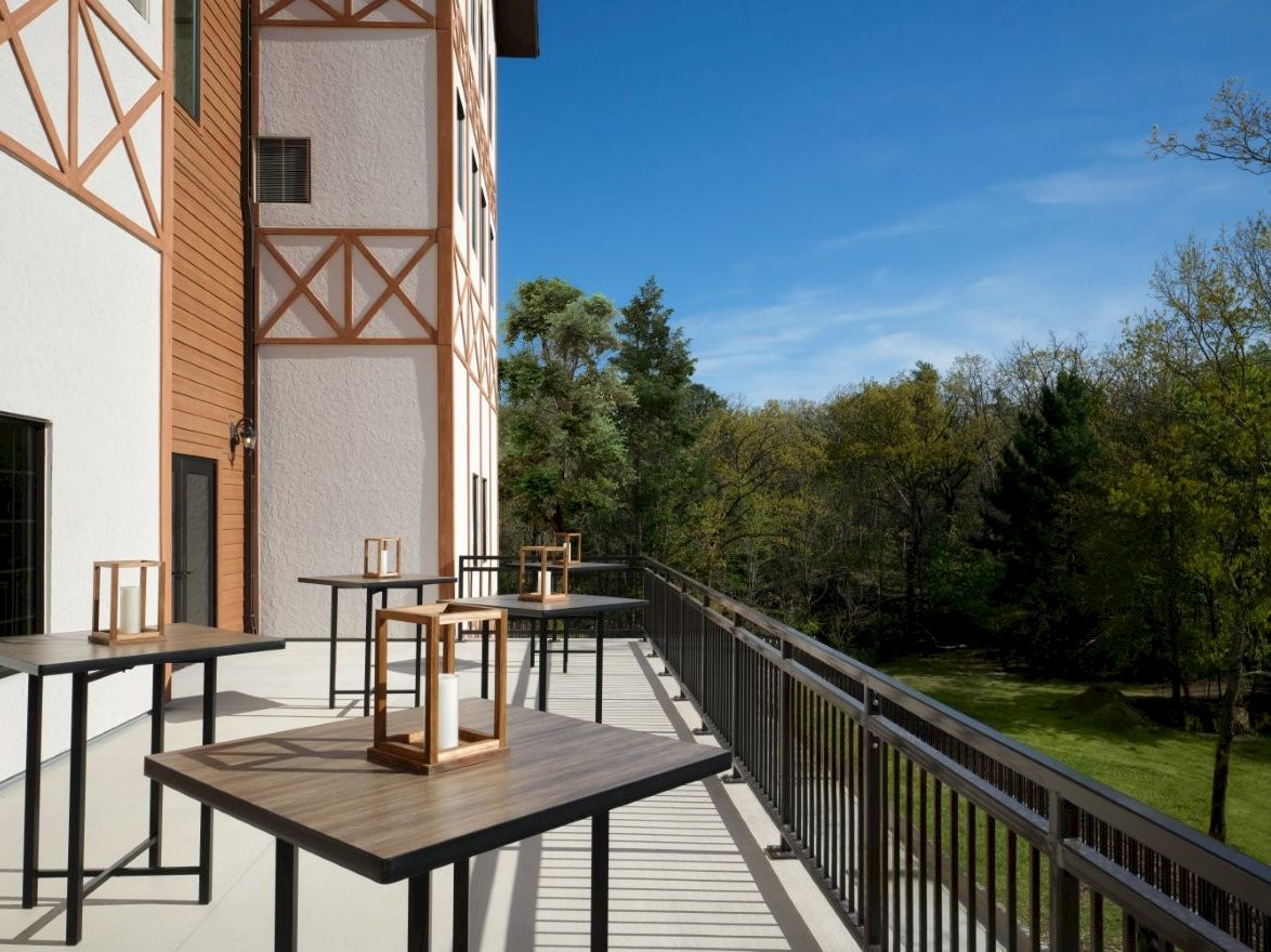 A sunny balcony with tables and lanterns, overlooking a green park and trees, attached to a brick building with wooden trim.