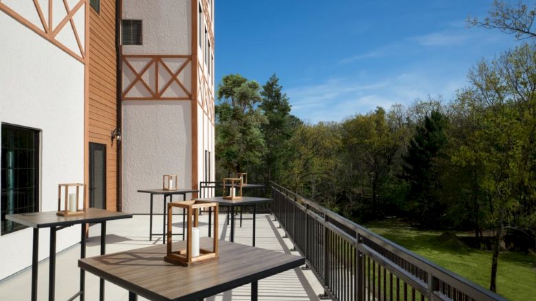 A sunny balcony with tables and lanterns, overlooking a green park and trees, attached to a brick building with wooden trim.