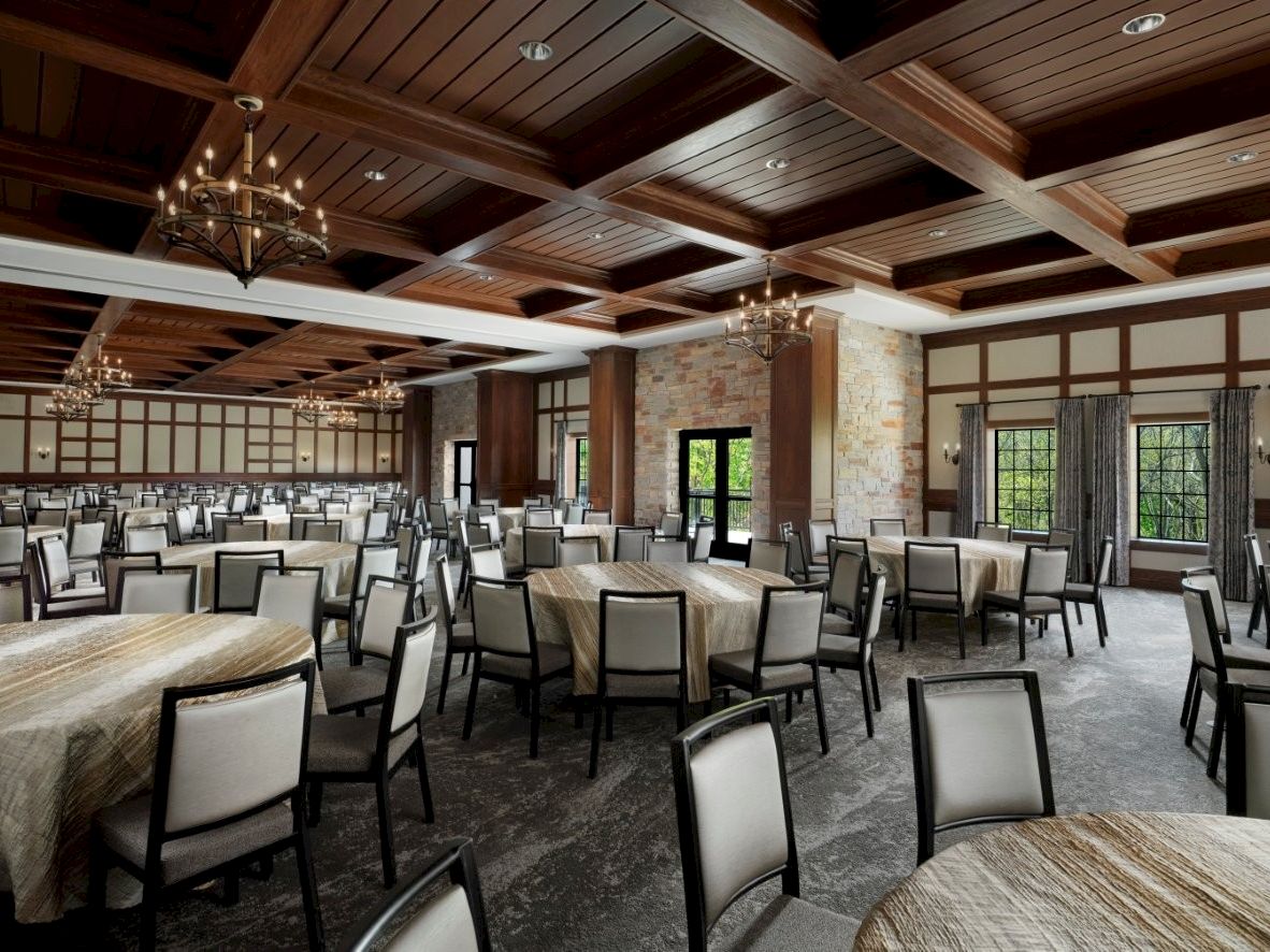 A spacious banquet hall with wood-beam ceiling, chandeliers, stone columns, and numerous round tables with padded chairs set for an event.