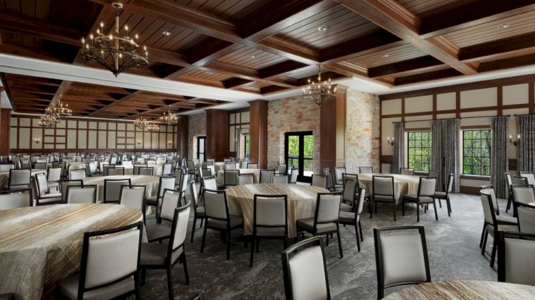 A spacious banquet hall with wood-beam ceiling, chandeliers, stone columns, and numerous round tables with padded chairs set for an event.