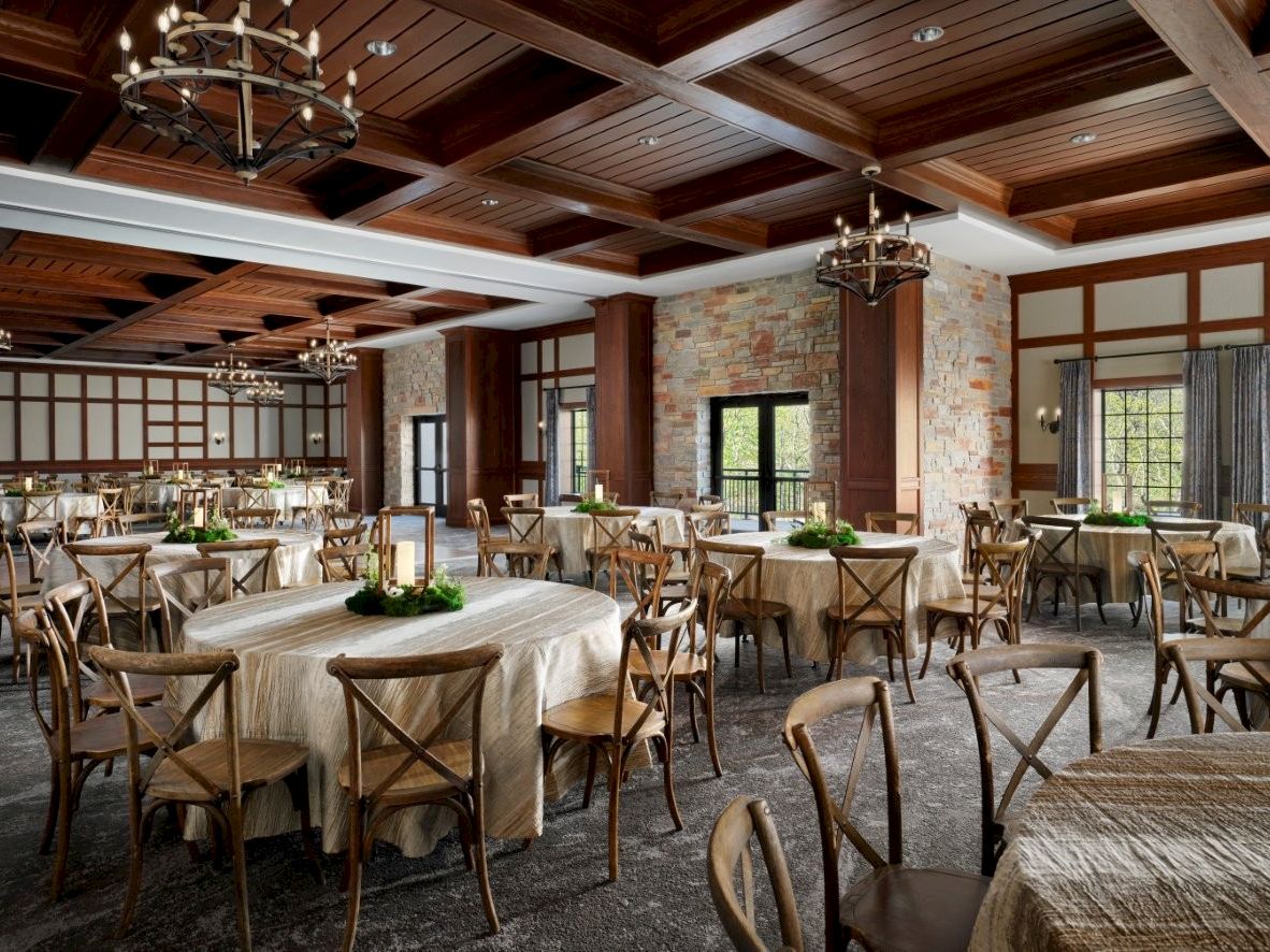 A spacious banquet hall with wooden beams, brick walls, chandeliers, and round tables set with linens and chairs, ready for a formal event.