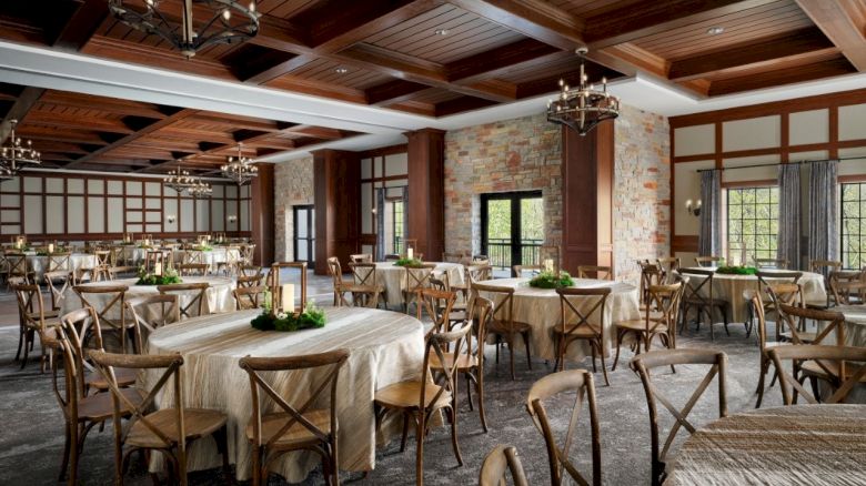 A spacious banquet hall with wooden beams, brick walls, chandeliers, and round tables set with linens and chairs, ready for a formal event.