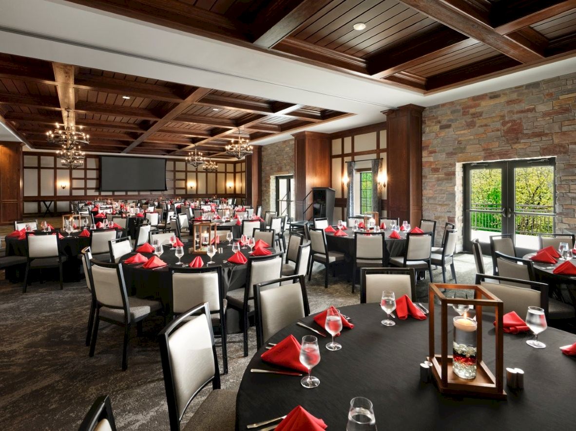 A banquet hall set for a formal event with round and rectangular tables, red napkins, white chairs, ambient lighting, brick walls, and large windows.