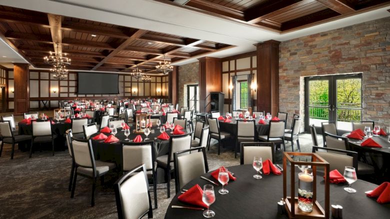 A banquet hall set for a formal event with round and rectangular tables, red napkins, white chairs, ambient lighting, brick walls, and large windows.