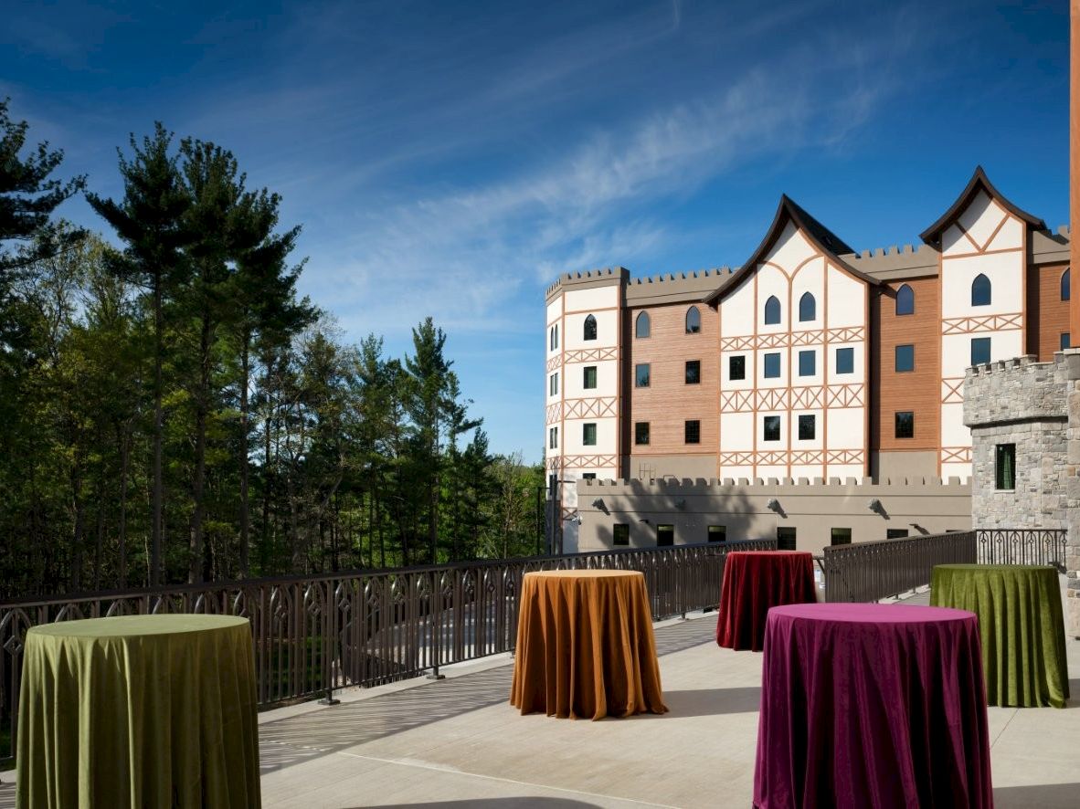 An outdoor terrace with round, colorfully covered cocktail tables in front of a castle-like building against a blue sky, surrounded by trees.