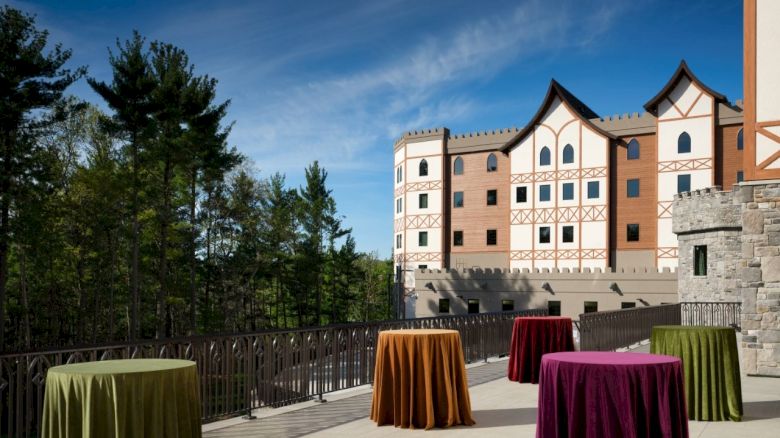 An outdoor terrace with round, colorfully covered cocktail tables in front of a castle-like building against a blue sky, surrounded by trees.
