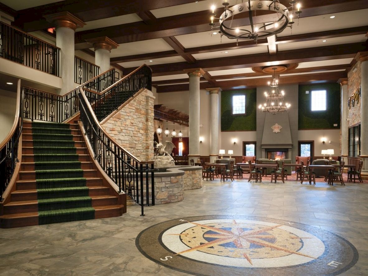 An elegant lobby with wooden staircases, green-carpeted steps, chandeliers, and a central seating area with fireplace.