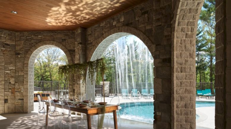 A stone-arched patio with a dining table, chairs, and a pool featuring cascading water fountains in the background.