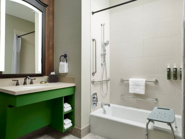 A modern bathroom with a green vanity, dark-framed mirror, white sink, and a tub/shower combo featuring accessibility rails and a shower chair, neat.