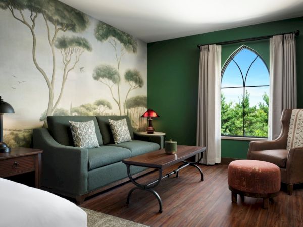 A cozy hotel room with a nature-mural wall, green accent wall, wooden floor, a sofa, coffee table, armchair with ottoman, and a large arched window letting in light.